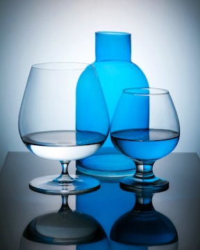 A composition of two glass goblets and a blue glass bottle. The goblets have reflections on a mirrored surface, and the bottle is positioned centrally in the background. The lighting casts soft shadows, highlighting the transparency and lines of the glassware.