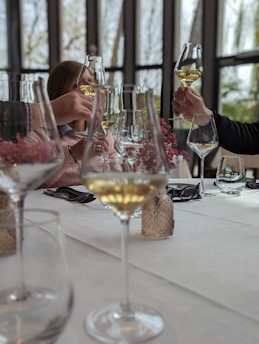 Hands holding glasses with white wine are raised in a toast around a table set with glassware and small vases of pink flowers. The setting appears to be indoors with a large window revealing trees in the background. The atmosphere suggests a social or celebratory gathering.