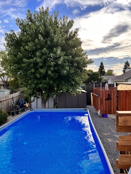 A backyard scene featuring a rectangular swimming pool with clear blue water. In the background, there is a large green tree with full foliage. A wooden fence encloses the area, and a shed is visible. The sky is partly cloudy with patches of blue visible.