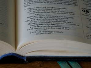 An open book displaying text on its pages. The pages are filled with printed words in a serif font, with one section of the text highlighted in blue ink. The image focuses on the text and shows the spine with a blue bookmark ribbon partially visible.