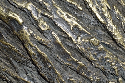 Metallic surface with an intricate pattern of silver and gold tones. The surface has a textured appearance with raised and indented sections, creating a rugged and dynamic visual effect.