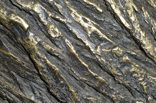 Metallic surface with an intricate pattern of silver and gold tones. The surface has a textured appearance with raised and indented sections, creating a rugged and dynamic visual effect.