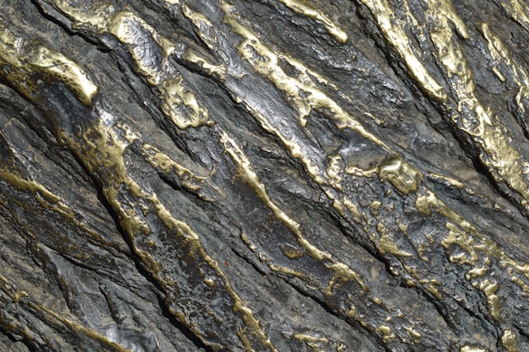 Metallic surface with an intricate pattern of silver and gold tones. The surface has a textured appearance with raised and indented sections, creating a rugged and dynamic visual effect.