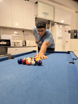 A person is preparing to break a set of billiard balls on a blue pool table. The surroundings are a kitchen area with white cabinets, appliances, and some signage on the wall.