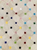 A pattern of beige tiles arranged in a geometric design, each tile featuring a small, brightly colored square in the corners. The squares vary in colors including blue, red, green, yellow, pink, and black, creating a visually appealing mosaic.
