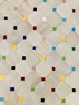 A pattern of beige tiles arranged in a geometric design, each tile featuring a small, brightly colored square in the corners. The squares vary in colors including blue, red, green, yellow, pink, and black, creating a visually appealing mosaic.