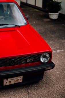A red vintage car is parked outdoors, showcasing its front hood and bumper. The car features a round headlight and a grill with a GTI emblem. The vehicle has a license plate from Paraguay, reading AUI-680. Potted plants are visible in the background.