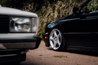 Two cars parked on a road with trees in the background; the focus is on the close-up of their wheels and bodywork.