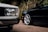 Two cars parked on a road with trees in the background; the focus is on the close-up of their wheels and bodywork.