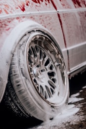 A vehicle tire and wheel are covered in thick soap suds, suggesting the car is undergoing a wash. The suds are dripping onto the ground, and the wheel design is intricate, with multiple spokes. The body of the car, visible in the background, is also coated in foam.