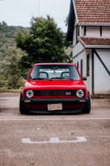 A classic red Volkswagen GTI is parked in front of a house with traditional architecture. The car has a vintage license plate from Paraguay and is parked on a concrete surface. Lush greenery and trees are visible in the background, adding a natural setting to the scene.