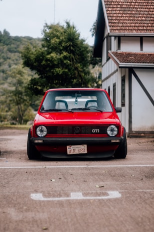 A classic red Volkswagen GTI is parked in front of a house with traditional architecture. The car has a vintage license plate from Paraguay and is parked on a concrete surface. Lush greenery and trees are visible in the background, adding a natural setting to the scene.