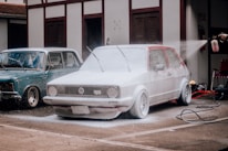 A red Volkswagen car is being washed with foam, covering most of its surface. Next to it is a blue vintage car parked in front of a building. A person is spraying water from a hose connected to a bottle, adding to the foam on the car. The setting appears to be an outdoor area with concrete pavement.
