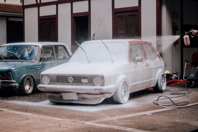 A red Volkswagen car is being washed with foam, covering most of its surface. Next to it is a blue vintage car parked in front of a building. A person is spraying water from a hose connected to a bottle, adding to the foam on the car. The setting appears to be an outdoor area with concrete pavement.