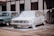 A red Volkswagen car is being washed with foam, covering most of its surface. Next to it is a blue vintage car parked in front of a building. A person is spraying water from a hose connected to a bottle, adding to the foam on the car. The setting appears to be an outdoor area with concrete pavement.