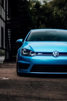 A vibrant blue car is parked on a street, with the front view dominating the image. The vehicle's glossy surface reflects the surrounding environment, and the car is positioned slightly off-center. A background of lush green foliage and a distant building are visible, creating a contrast with the car's color.