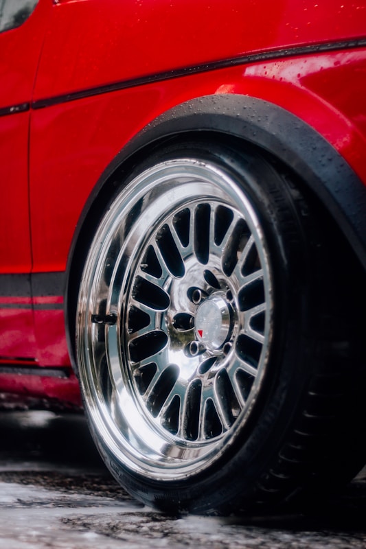 A close-up view of a shiny chrome car wheel with a detailed rim design, attached to a red vehicle. Water droplets are visible on the car's body and the tire, suggesting recent rain or washing.
