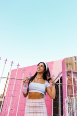 A woman with long dark hair is standing in front of a pink building, holding a bottle of Jarritos soda. She is wearing a white crop top, a plaid skirt, and has various bracelets on her wrist. She is smiling and looking off into the distance. The background includes a clear blue sky and a decorative metal fence with pink accents.