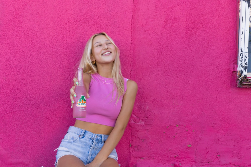 a woman sitting against a pink wall holding a bottle of water