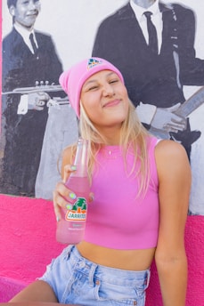 A woman wearing a pink beanie and pink sleeveless top is holding a bottle of Jarritos guava-flavored soda. She is smiling with a playful expression, and is sitting in front of a mural depicting two men in suits playing musical instruments.