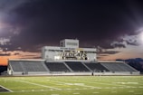 A football stadium with empty bleachers and a large central building displaying the words 'Wayne State College Wildcats'. The sky is moody with dark clouds, suggesting a sunset or impending storm in the background.