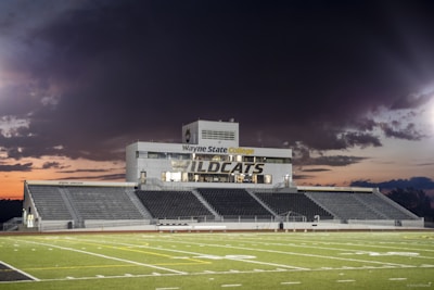 A football stadium with empty bleachers and a large central building displaying the words 'Wayne State College Wildcats'. The sky is moody with dark clouds, suggesting a sunset or impending storm in the background.