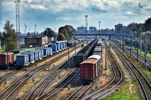 A railway yard featuring multiple tracks with several freight trains carrying various colored containers. The scene is set in an industrial background with buildings, utility poles, and trees lining the sides.