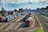 A railway yard featuring multiple tracks with several freight trains carrying various colored containers. The scene is set in an industrial background with buildings, utility poles, and trees lining the sides.