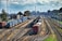 A railway yard featuring multiple tracks with several freight trains carrying various colored containers. The scene is set in an industrial background with buildings, utility poles, and trees lining the sides.
