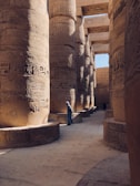 Massive ancient stone pillars adorned with intricate hieroglyphics create a majestic corridor. A person in traditional attire walks along the path, highlighting the scale of the architectural marvel. Sunlight casts gentle shadows, enhancing the texture of the carvings.