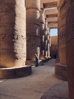 Massive ancient stone pillars adorned with intricate hieroglyphics create a majestic corridor. A person in traditional attire walks along the path, highlighting the scale of the architectural marvel. Sunlight casts gentle shadows, enhancing the texture of the carvings.