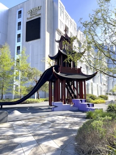 A pagoda-style playground structure with a slide is set amidst a landscaped area with trees and shrubs. Modern buildings are visible in the background, and the scene is well lit with natural sunlight.