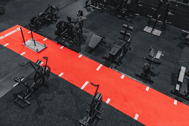 A gym setting featuring a variety of exercise equipment such as weight machines, stationary bikes, and a sled on a bright red track. The gym has a modern aesthetic with black and red color schemes.