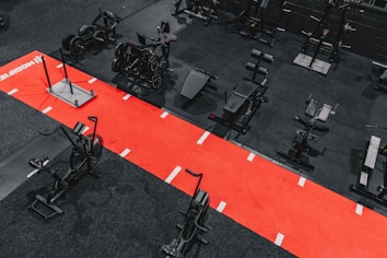 A gym setting featuring a variety of exercise equipment such as weight machines, stationary bikes, and a sled on a bright red track. The gym has a modern aesthetic with black and red color schemes.