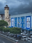 A blue building with white pillars identified as the Myanma Tourism Bank is located on a busy city street. Several cars are parked near the sidewalk along the road, and people are walking nearby. The sky above the scene is cloudy and overcast. An old, tall, beige clock tower with a flag is visible next to the bank building. Trees line the street, adding greenery to the urban environment.