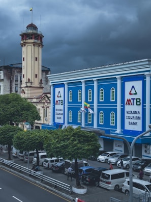 A blue building with white pillars identified as the Myanma Tourism Bank is located on a busy city street. Several cars are parked near the sidewalk along the road, and people are walking nearby. The sky above the scene is cloudy and overcast. An old, tall, beige clock tower with a flag is visible next to the bank building. Trees line the street, adding greenery to the urban environment.