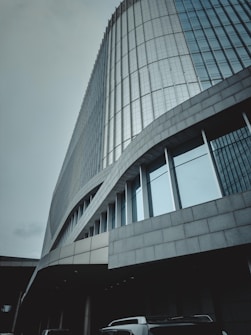 A modern, sleek skyscraper with a curved facade covered in reflective glass panels. The building's architecture features smooth lines and a combination of stone and glass materials. Several vehicles, including what appears to be vans, are positioned near the entrance at the lower level.