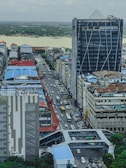 An urban landscape featuring a busy street with a line of cars visible between tall buildings. The foreground has a prominent building with signage for KBZ Bank. In the background, a river with a lush green forest on the far bank can be seen under an overcast sky.