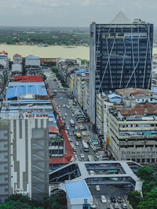 An urban landscape featuring a busy street with a line of cars visible between tall buildings. The foreground has a prominent building with signage for KBZ Bank. In the background, a river with a lush green forest on the far bank can be seen under an overcast sky.