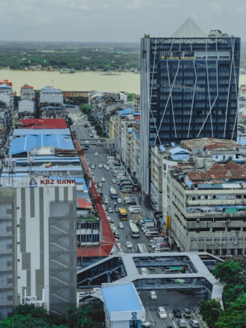 An urban landscape featuring a busy street with a line of cars visible between tall buildings. The foreground has a prominent building with signage for KBZ Bank. In the background, a river with a lush green forest on the far bank can be seen under an overcast sky.