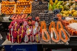 A variety of fresh fruits neatly displayed at a market stall. There are halved papayas with black seeds visible, dragon fruits sliced in half with white flesh and black seeds, and yellow spoons inserted into them. Nearby, there are containers with dates and other exotic fruits such as melons and oranges arranged in stacks.