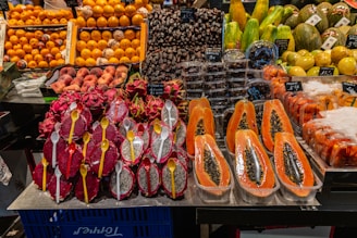 A variety of fresh fruits neatly displayed at a market stall. There are halved papayas with black seeds visible, dragon fruits sliced in half with white flesh and black seeds, and yellow spoons inserted into them. Nearby, there are containers with dates and other exotic fruits such as melons and oranges arranged in stacks.