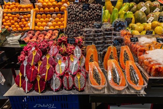 A variety of fresh fruits neatly displayed at a market stall. There are halved papayas with black seeds visible, dragon fruits sliced in half with white flesh and black seeds, and yellow spoons inserted into them. Nearby, there are containers with dates and other exotic fruits such as melons and oranges arranged in stacks.