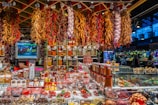 A vibrant market stall is filled with an array of spices, herbs, and produce. Bundles of dried chili peppers and garlic hang from the ceiling, while shelves are stocked with various containers of spices. The stall has a colorful assortment of products, and a small television displays images of fresh produce. The atmosphere is bustling, with shoppers visible in the background.