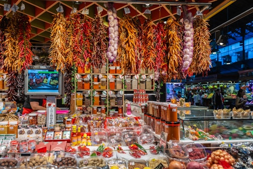 A vibrant market stall is filled with an array of spices, herbs, and produce. Bundles of dried chili peppers and garlic hang from the ceiling, while shelves are stocked with various containers of spices. The stall has a colorful assortment of products, and a small television displays images of fresh produce. The atmosphere is bustling, with shoppers visible in the background.