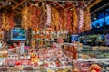 A vibrant market stall is filled with an array of spices, herbs, and produce. Bundles of dried chili peppers and garlic hang from the ceiling, while shelves are stocked with various containers of spices. The stall has a colorful assortment of products, and a small television displays images of fresh produce. The atmosphere is bustling, with shoppers visible in the background.