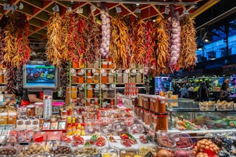 A vibrant market stall is filled with an array of spices, herbs, and produce. Bundles of dried chili peppers and garlic hang from the ceiling, while shelves are stocked with various containers of spices. The stall has a colorful assortment of products, and a small television displays images of fresh produce. The atmosphere is bustling, with shoppers visible in the background.