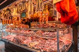 A vibrant meat market stall displaying a wide variety of cured meats and sausages. Different cuts of meat hang from the top, while an assortment of packaged meat products are neatly arranged in the glass display case below. The stall is warmly lit, creating an inviting atmosphere for shoppers.