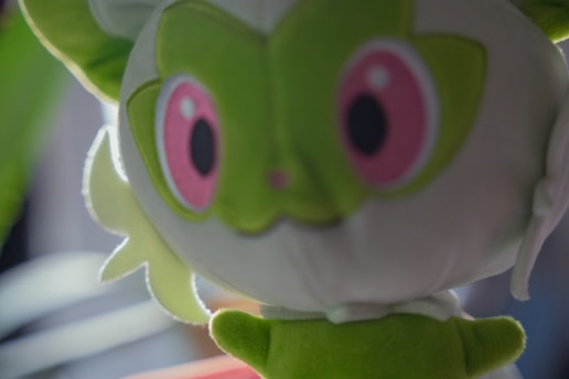 A plush toy with large pink eyes and lime green facial features. It has a whimsical and cute appearance, with fluffy ears and a small mouth.