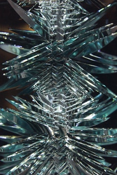 A symmetrical arrangement of overlapping translucent glass-like blades, creating a visually intricate and reflective pattern. The design appears dynamic and three-dimensional, with a combination of sharp angles and smooth surfaces.
